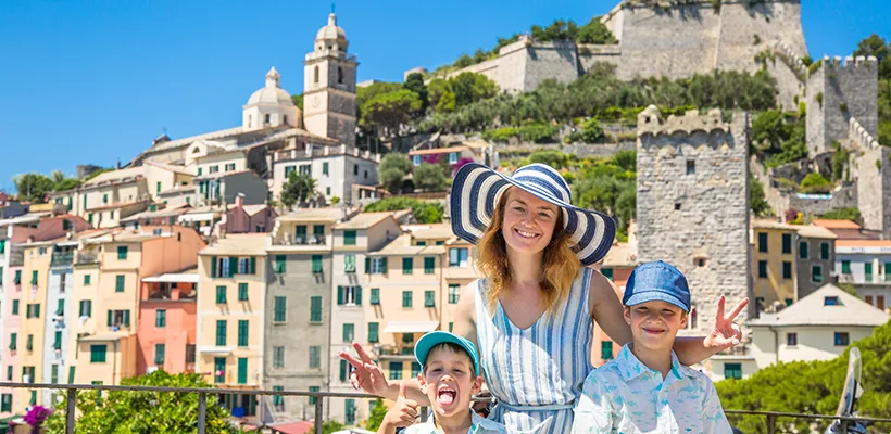 Une maman avec ses deux fils posant devant un village des cinque terre en Italie. La maman porte un chapeau rayé blanc et bleu faisant deux signes « peace » avec ses doigts,  un enfant fait un pouce avec ses doigts pendant que l’autre enfant plus à sa gauche sourit 