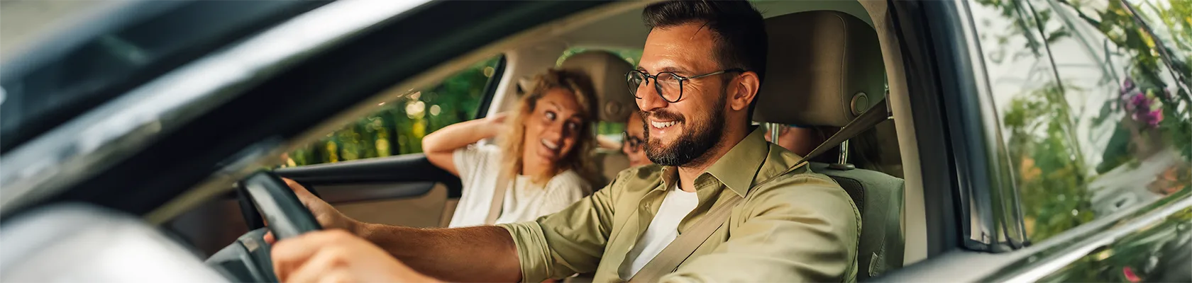 Départ en vacances d’une famille dans une voiture avec le papa qui conduit le sourire aux lèvres, la maman qui regarde vers l’arrière de la voiture s’adressant à ses enfants.